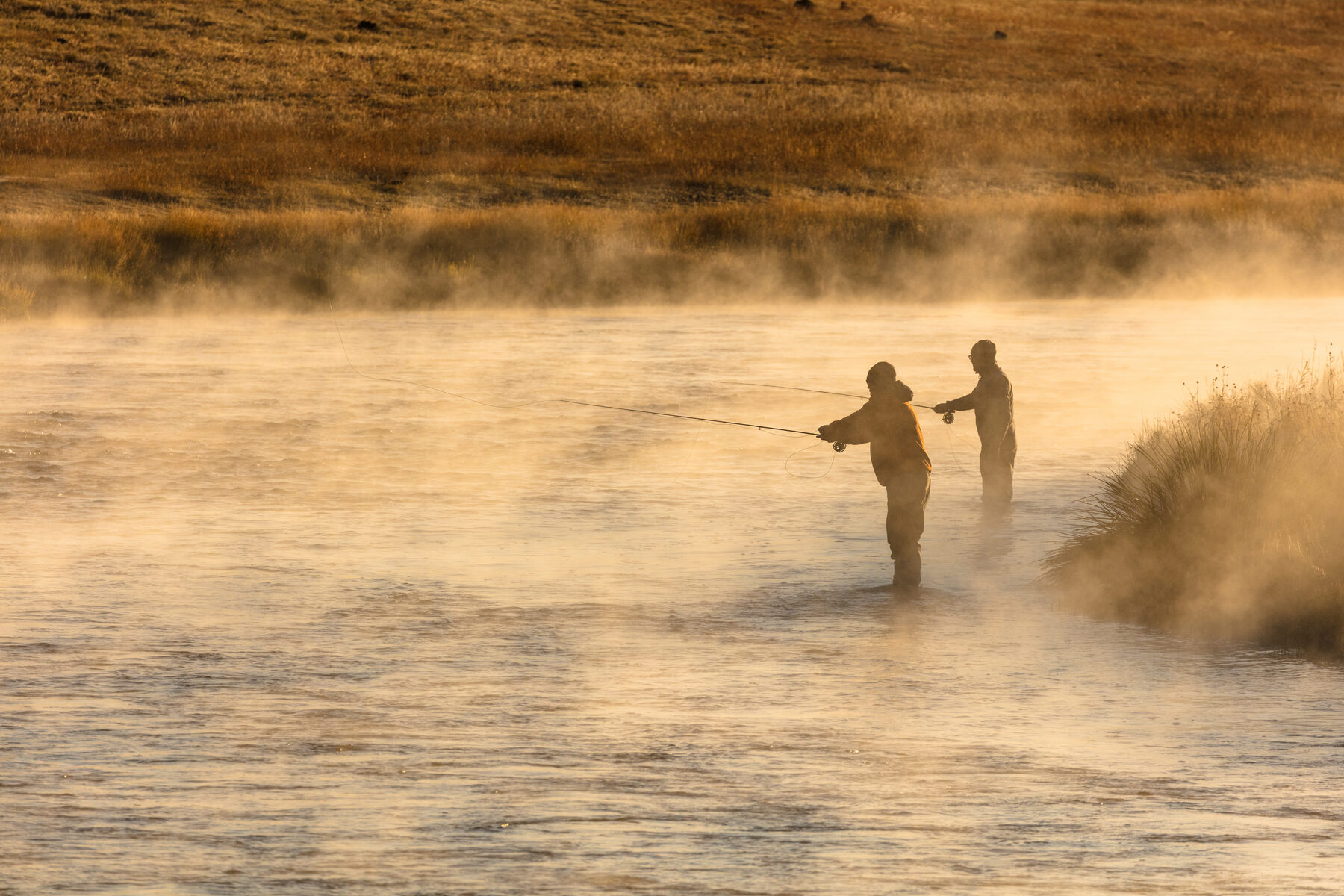 Two anglers fly fishing at sunrise with golden mist rising off the river